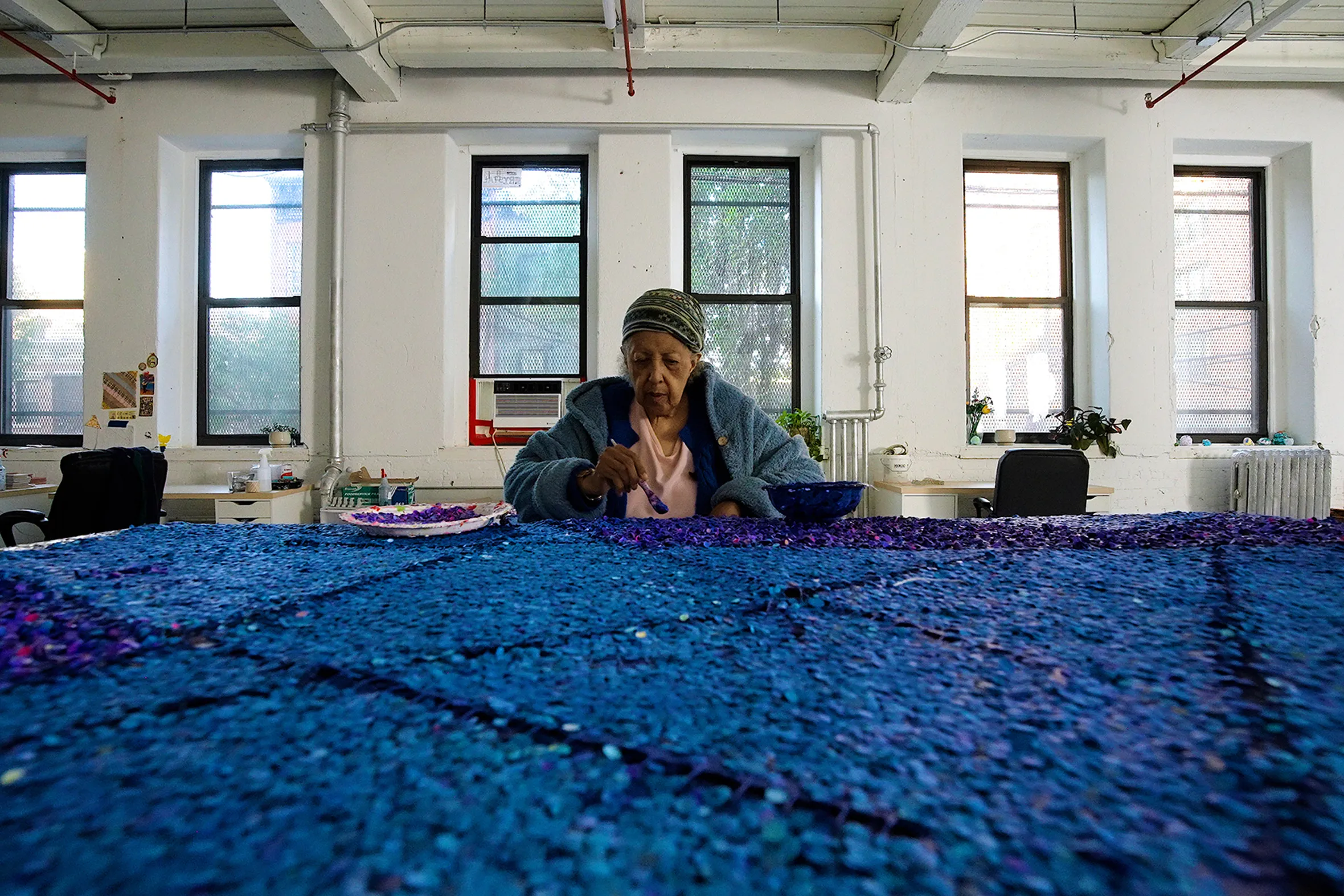 An elderly woman sits at a table covered in blue material, working on a project in a brightly lit studio with windows.