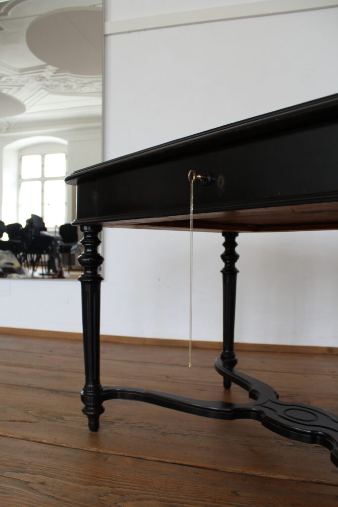 Black ornate desk with a key hanging from the drawer on a wooden floor.