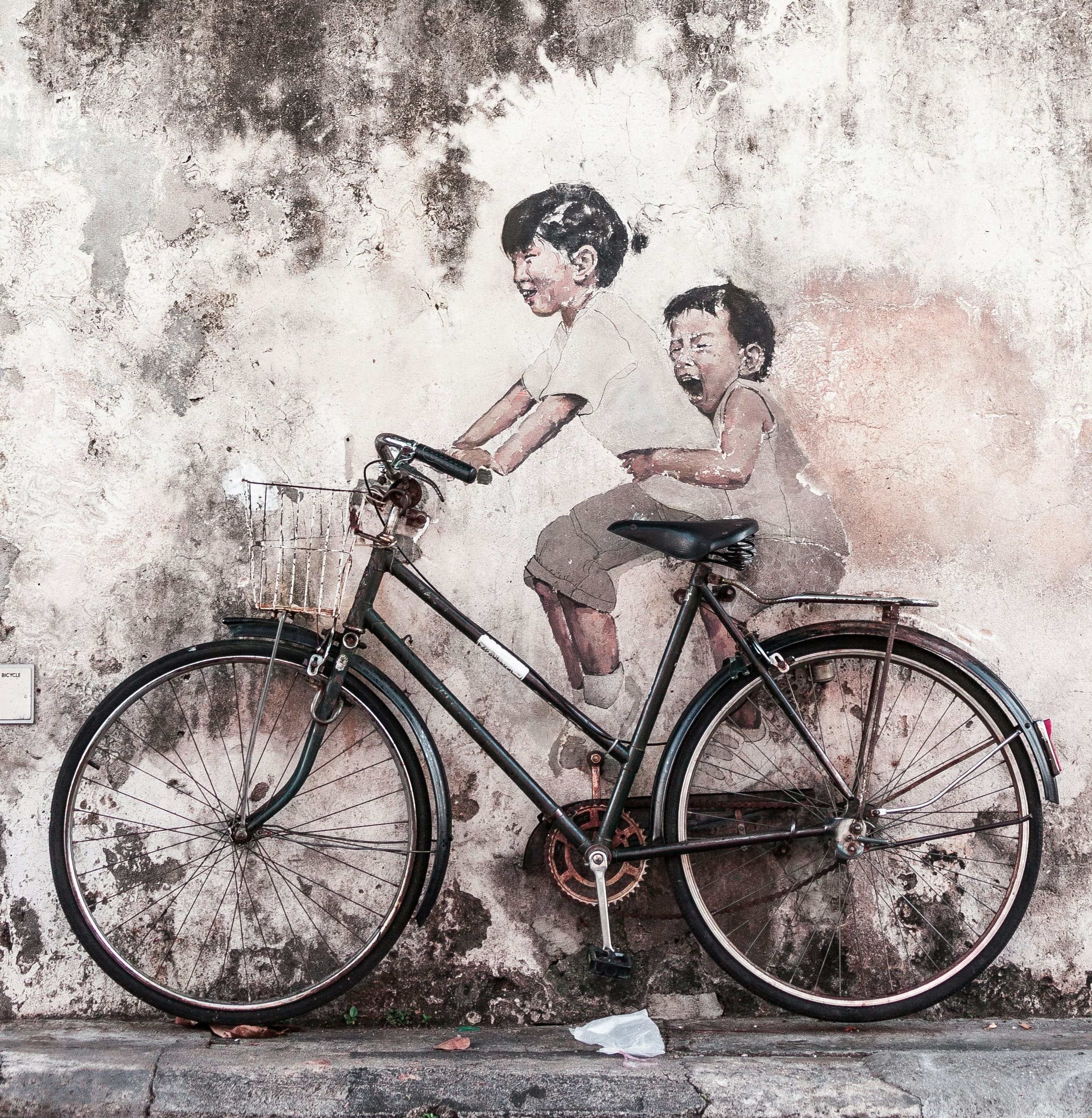 A vintage bicycle leans against a weathered wall featuring a faded mural of two children playfully riding together. | Photo by Yaopey Yong on Unsplash