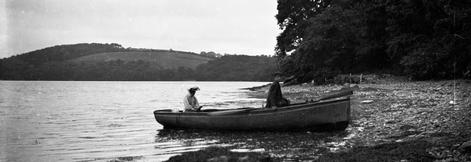 A man and a woman sit in a small boat on a calm river, surrounded by lush hills and wooded shorelines under an overcast sky.