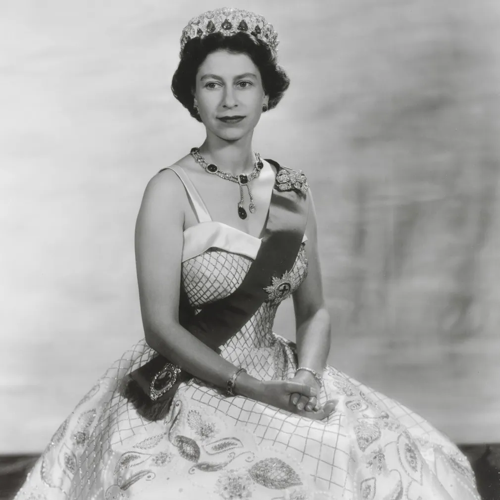 A woman elegantly dressed in an intricately patterned ball gown, adorned with jewels, seated gracefully against a soft backdrop.