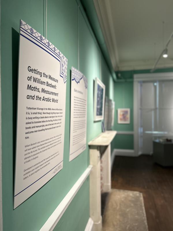 A room featuring several signs on the wall, including a whiteboard. The space is part of an exhibition titled "Getting the Measure of William Bedwel: Maths, Measurement and the Arabic Word." The ceiling and floor are visible in the indoor setting.
