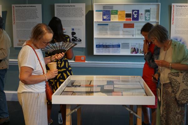 A group of people standing around a table indoors, with various clothing styles visible. The table is surrounded by furniture and there are artworks or designs on the wall in the background. | 2025 Bruce Castle Museum