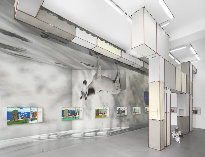 A contemporary art installation featuring a large cow mural, hanging structures, and colorful framed artworks on display.