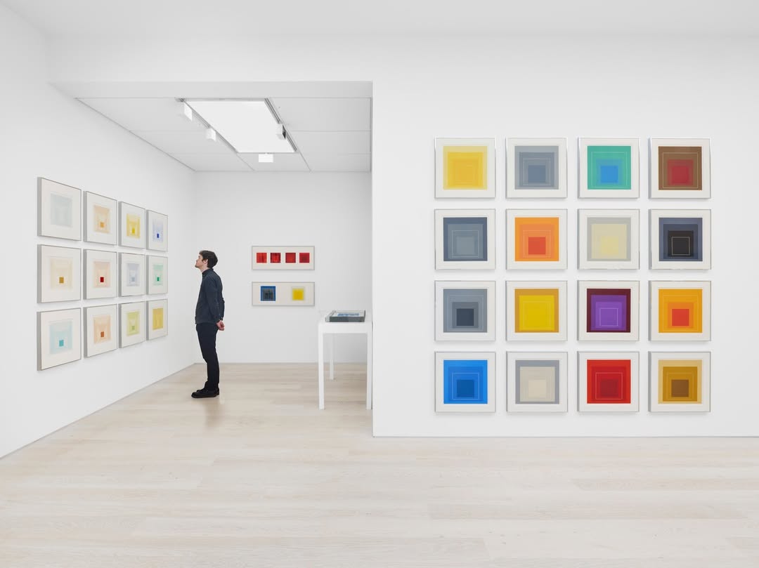 A visitor admires abstract square art pieces on white walls in a modern gallery, highlighting vibrant colors and geometric designs.