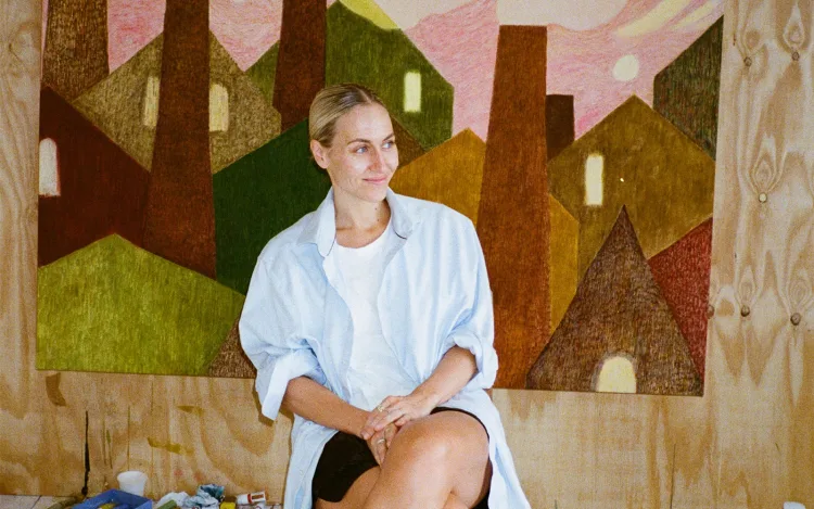 A person sits casually in front of a colorful mural depicting abstract houses and landscapes, exuding a creative atmosphere.