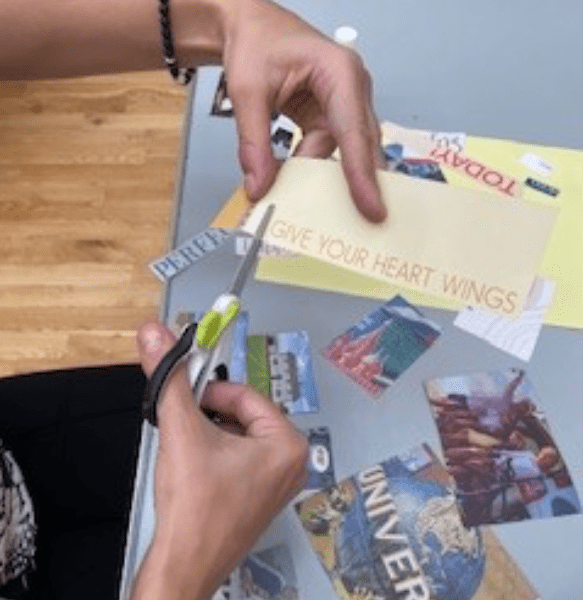 A hand holds scissors, cutting out a yellow paper with the text "GIVE YOUR HEART WINGS," surrounded by colorful magazine clippings.