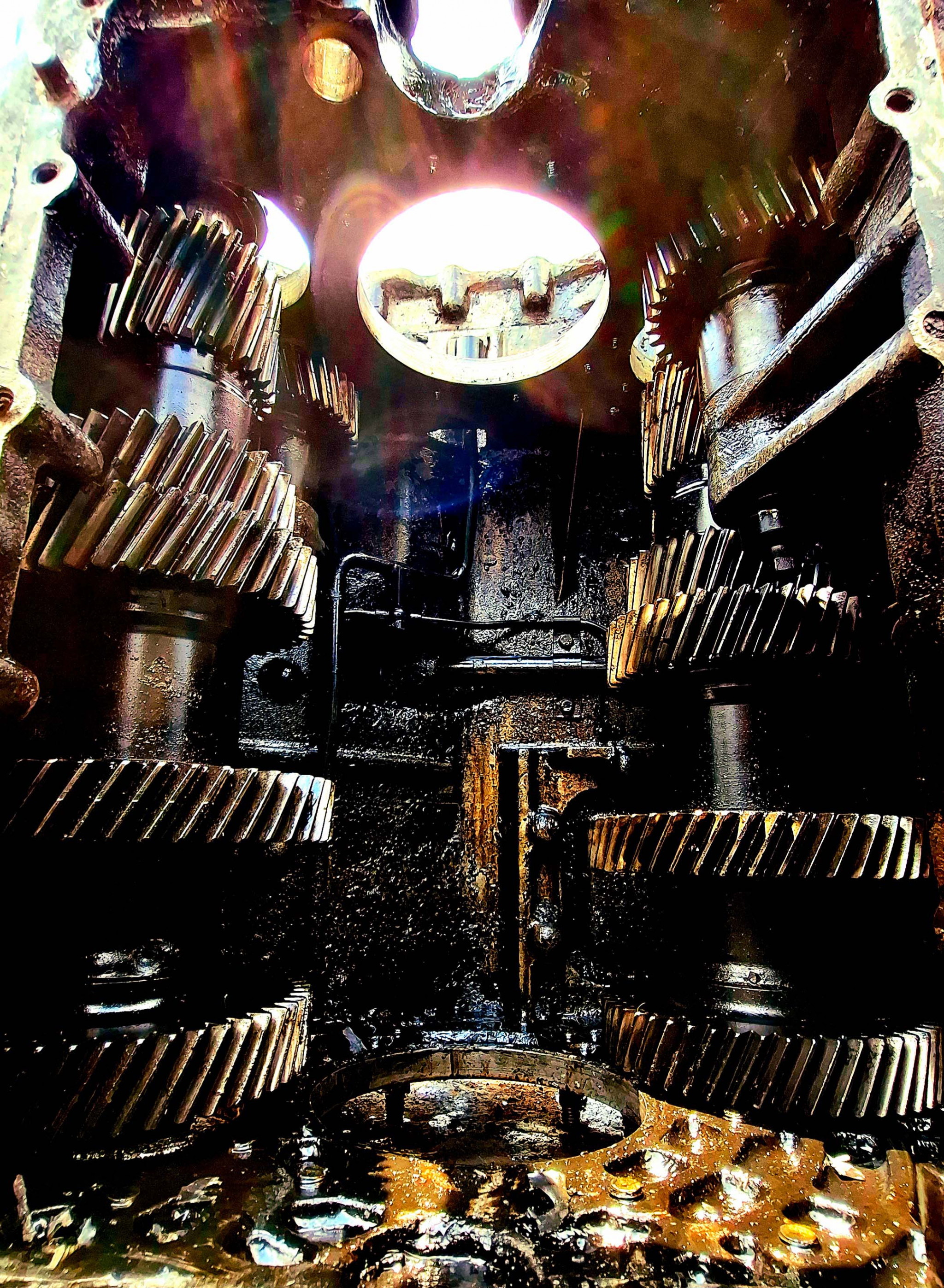 Interior view of a complex gear system, showing interlocking metallic gears, with oil residue and textured surfaces. Light enters from above.