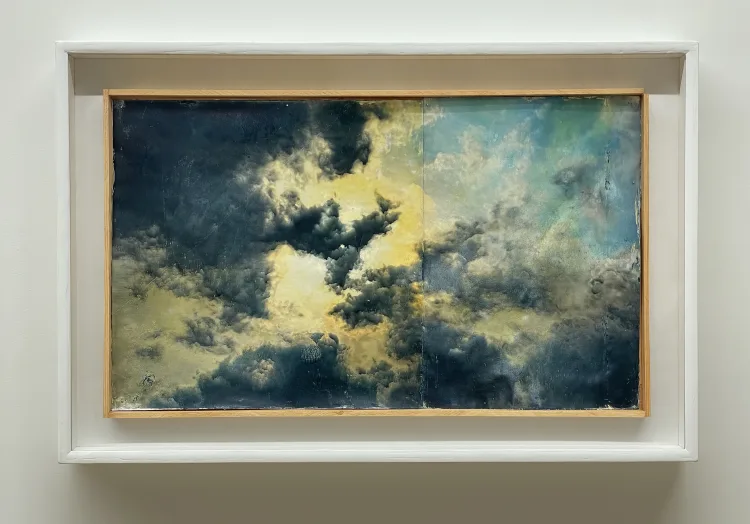 A painting depicting a cloudy sky, displayed in a white frame within an art gallery setting.