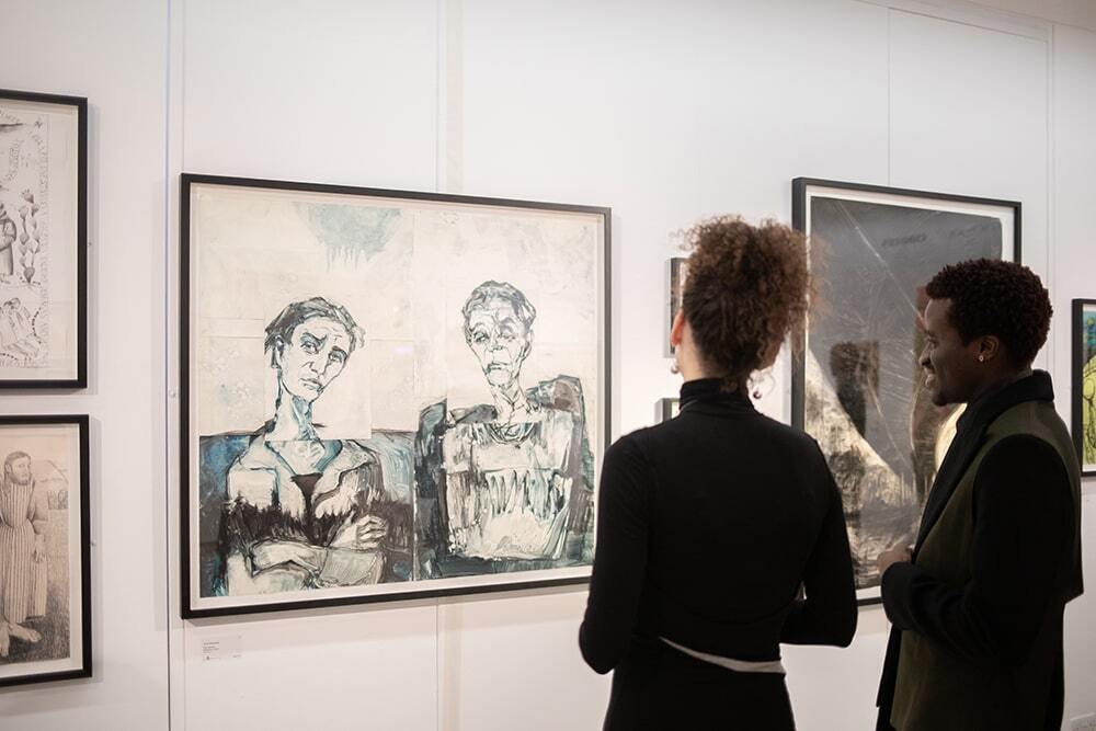 A group of people standing in an indoor art gallery, observing various paintings displayed on a wall within a room.