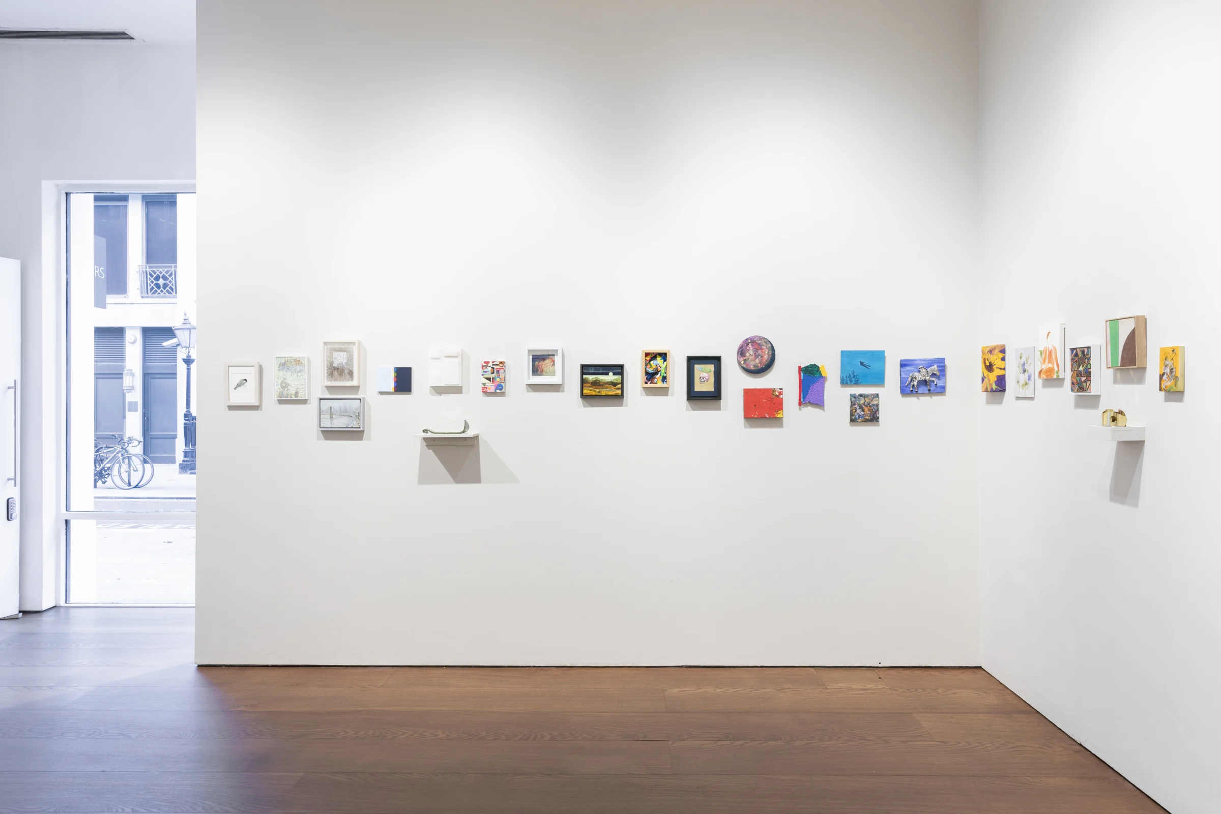 A room featuring a white wall adorned with various pictures, showcasing an indoor art gallery atmosphere. The floor is visible, contributing to the contemporary feel of the space.