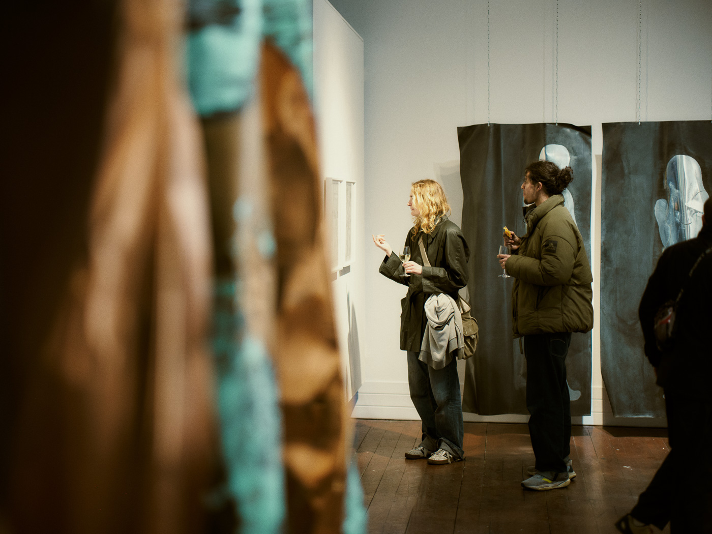 Two individuals observe art in a gallery, surrounded by colorful artworks and muted wall pieces, holding drinks and snacks.