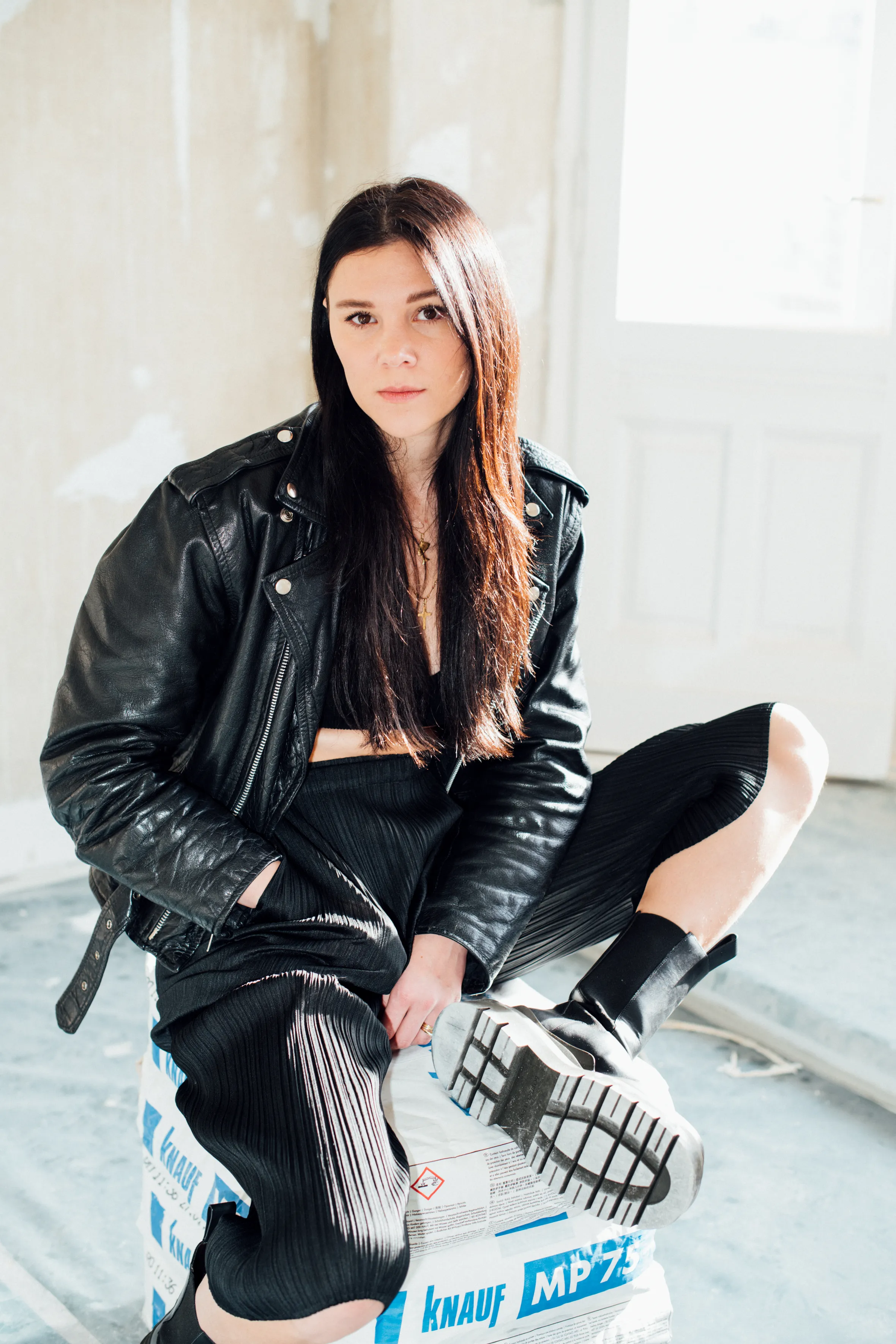 A woman with long hair wearing a leather jacket poses indoors against a wall, showcasing her fashion style.