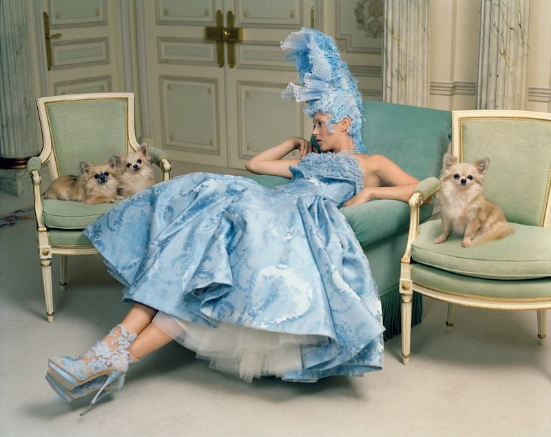 A woman in a blue dress is lying on a green chair with small dogs around her. The setting is indoors, featuring furniture and a wall in the background.
