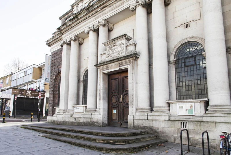 A classical government building featuring a prominent door and steps, with columns adorning the facade. The structure is made of stone, set against a clear sky in a city environment during winter. A statue can be seen nearby, adding to the architectural details of the outdoor scene.