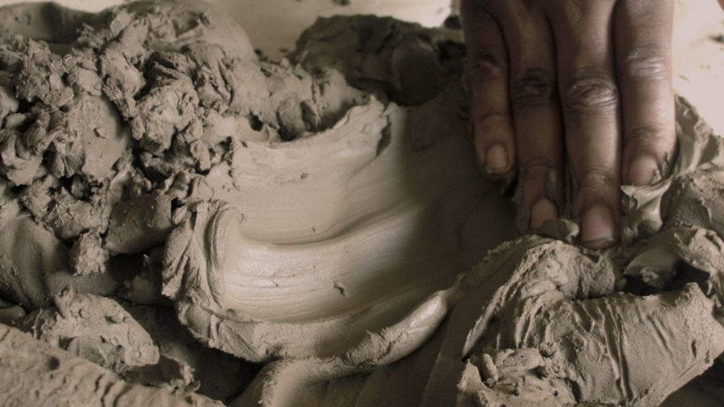 A close-up of a person's hand shaping clay on a pottery wheel, with a statue and vase in the background. The image is in black and white.