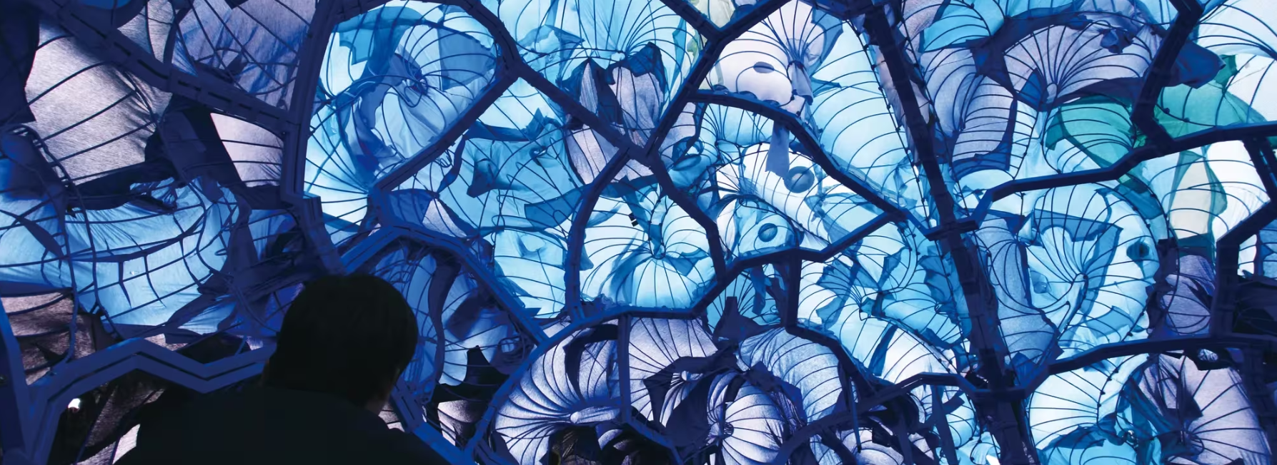 A viewer gazes up at a captivating blue and turquoise illuminated installation featuring intricate fabric shapes resembling petals and leaves. | 2025 Hayward Gallery