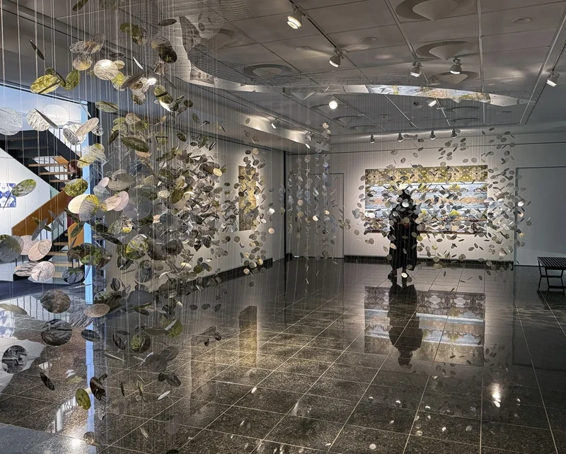 A person stands among hanging leaf-like artworks in a bright gallery, with textured paintings visible on the walls.