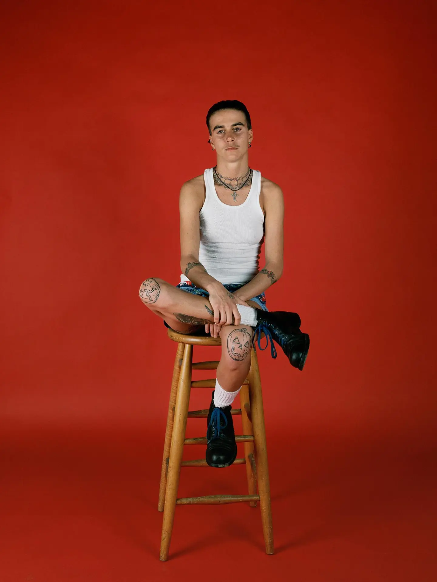 A man sitting on a stool indoors, against a wall, wearing various clothing and footwear. The setting includes furniture and showcases human features.