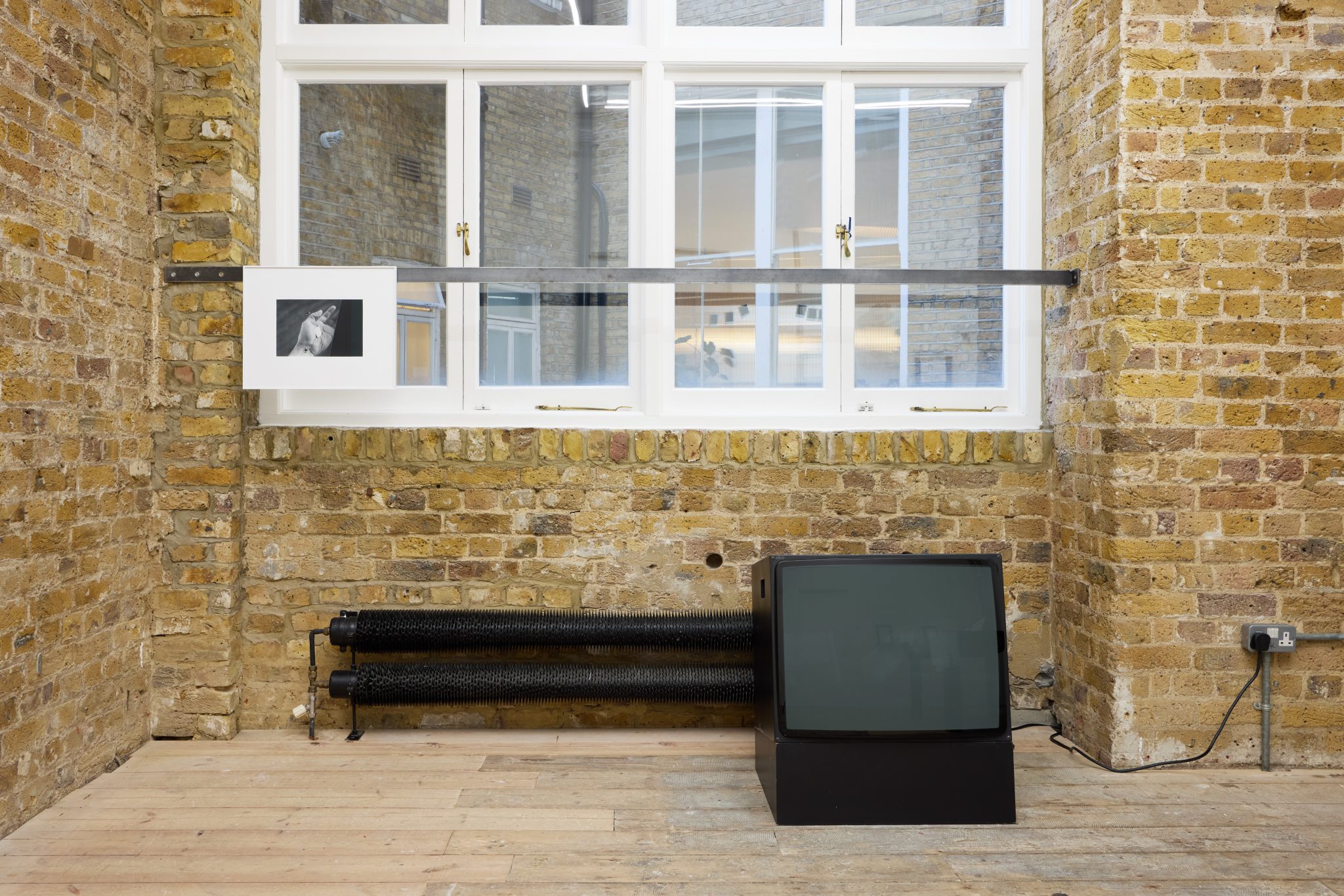 A television is positioned in a room with a window, showcasing elements like brick walls and a bench, indicating an indoor setting that opens to the outdoors.