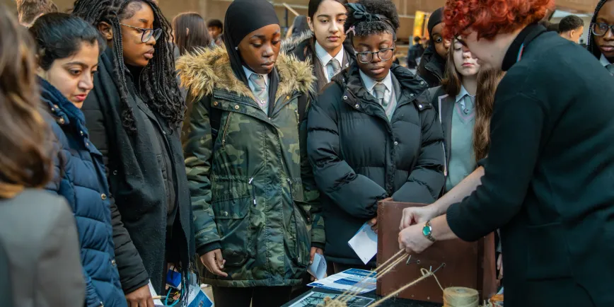 A diverse group of people standing together in a crowd, with some wearing jackets and suits. They are smiling and engaging with each other in an outdoor setting.