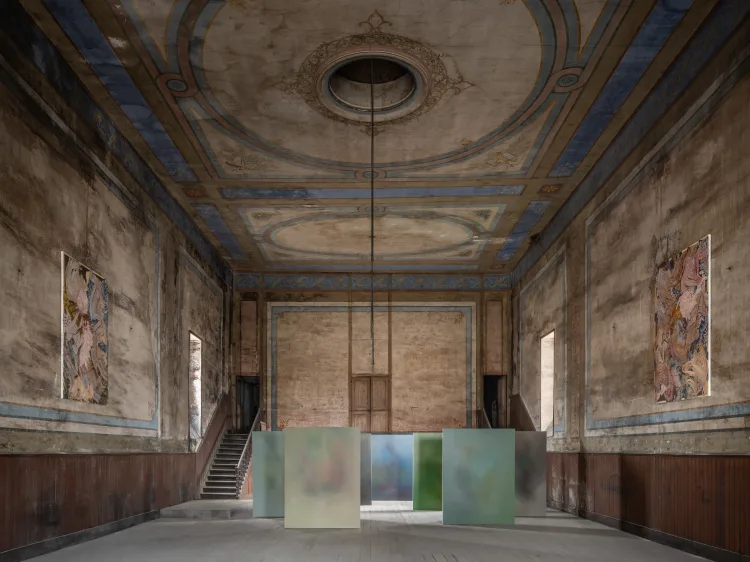 A spacious abandoned room featuring a deteriorating ceiling and a staircase leading to the upper level. The walls show signs of decay with peeling plaster, and remnants of art can be seen in various places on the floor.