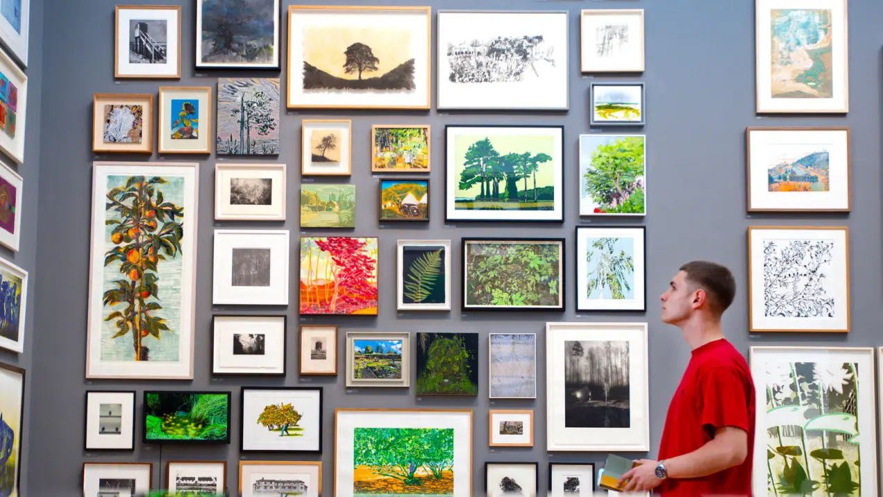 A person in a red shirt observes a gallery wall displaying numerous framed artworks, mostly nature-themed paintings and photographs, arranged in a grid pattern.