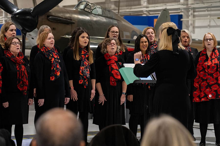 A choir dressed in black dresses and red poppy scarves performs in front of a vintage aircraft, with a conductor leading them.