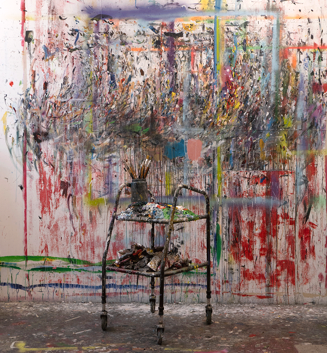 An artist's cart with brushes and paint sits in front of a vibrant, paint-splattered wall, showcasing creative chaos.