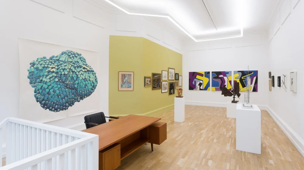 A room featuring a desk against the wall, adorned with various pieces of art displayed in frames. The floor is visible, and the overall interior design gives a gallery-like atmosphere.