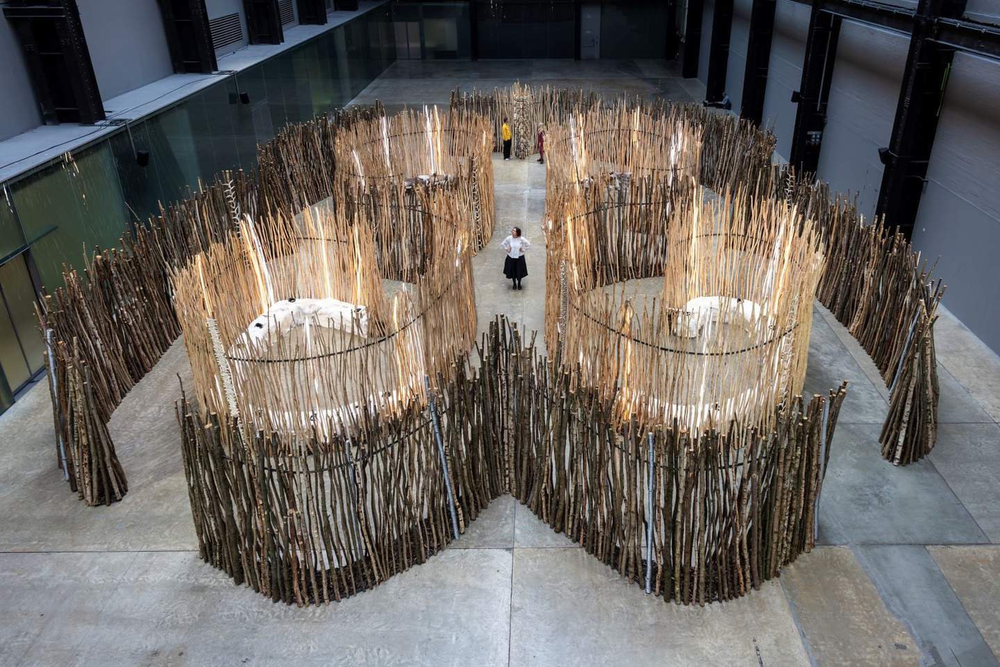 A group of people standing in a room with a large structure made of sticks, surrounded by furniture and art on the floor.