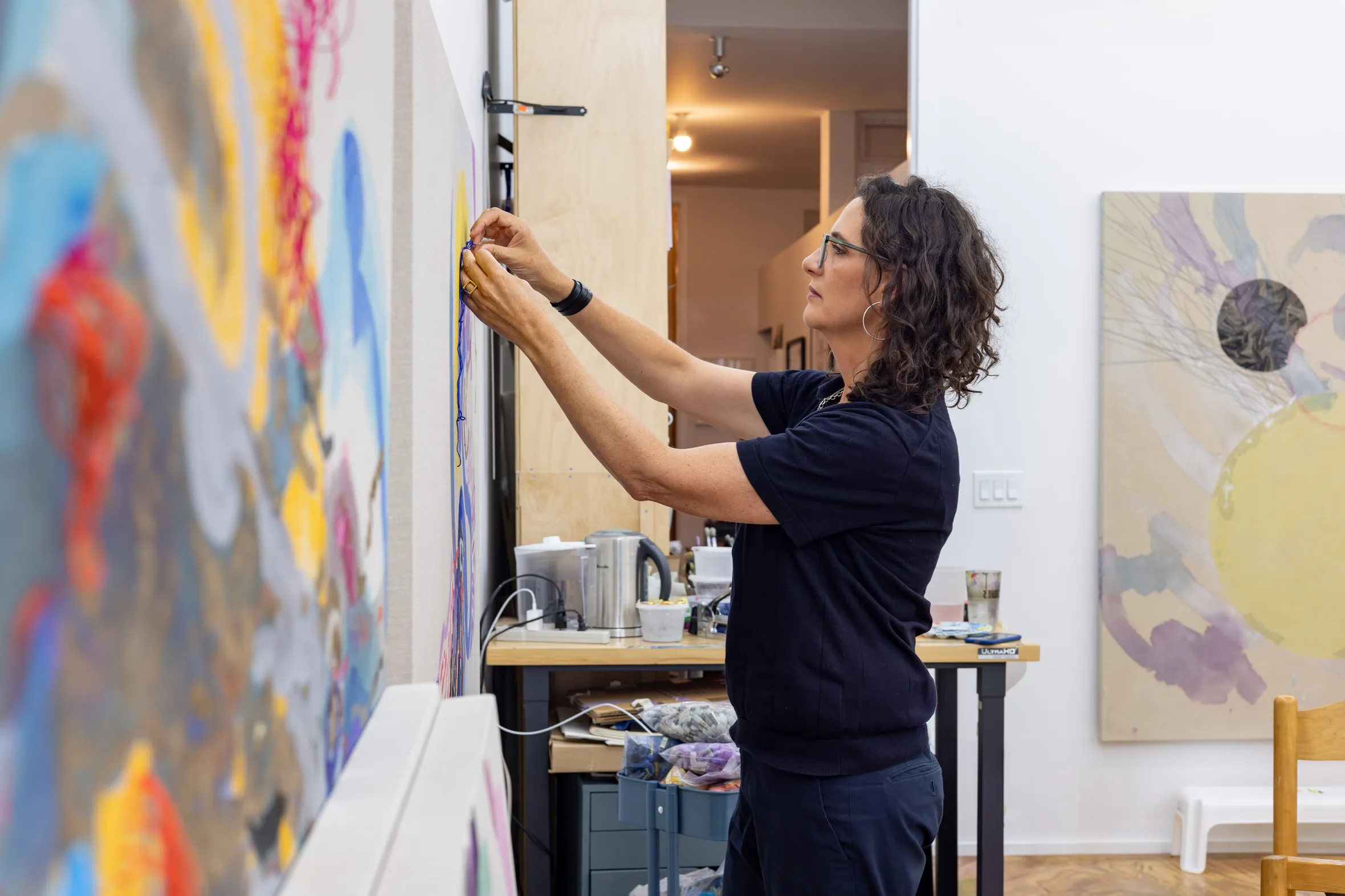 A woman is painting a wall indoors, surrounded by furniture including a chair and a table. There is a whiteboard nearby and a tripod set up for support. She is wearing casual clothing while focused on her artwork.