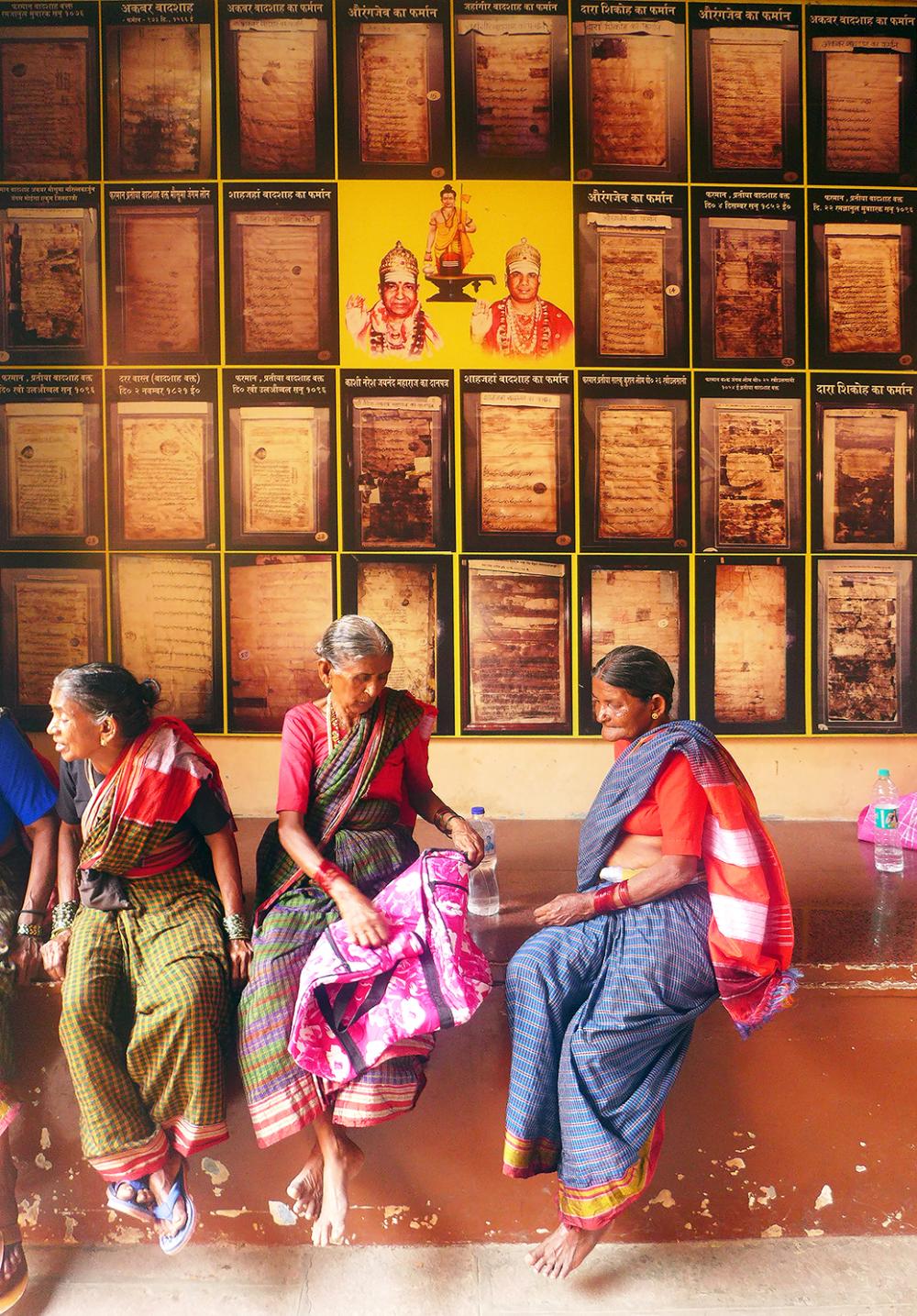 A group of women sitting on a wall, surrounded by artworks and pictures. They are dressed in various clothing styles, showcasing different footwear. The setting appears to be indoors with a floor beneath them.