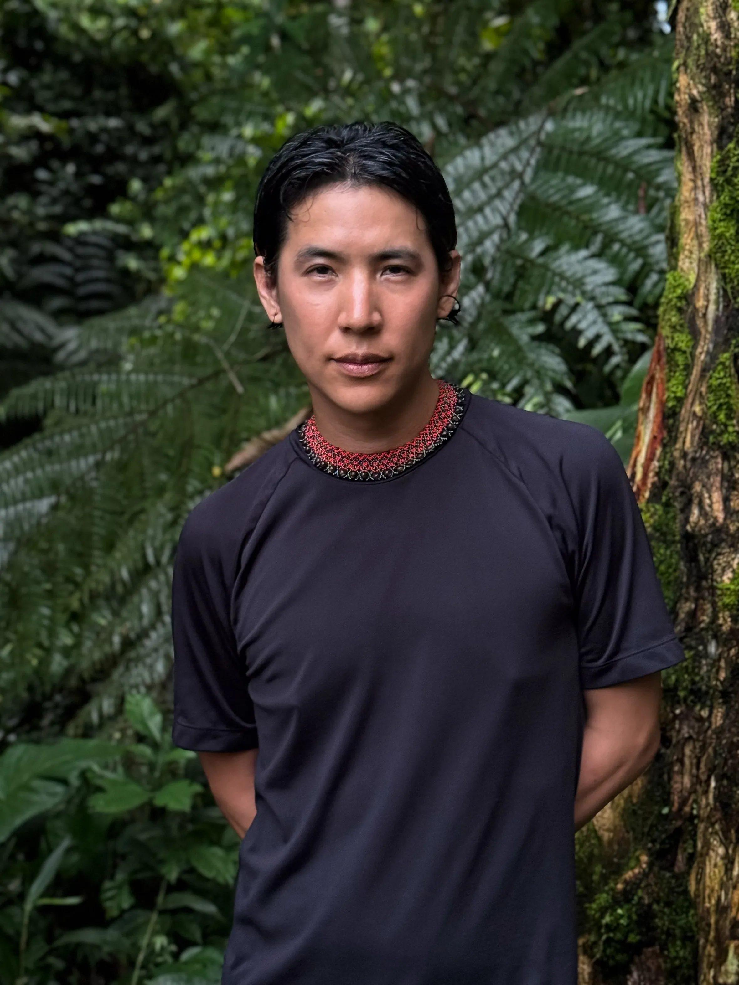 A man is standing in front of a large tree, smiling. He is outdoors, surrounded by a forest with lush greenery. The man is wearing a shirt and appears to be enjoying a hiking experience.