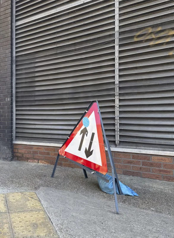A road sign positioned on the sidewalk, with a building in the background, surrounded by a traffic cone on the ground.