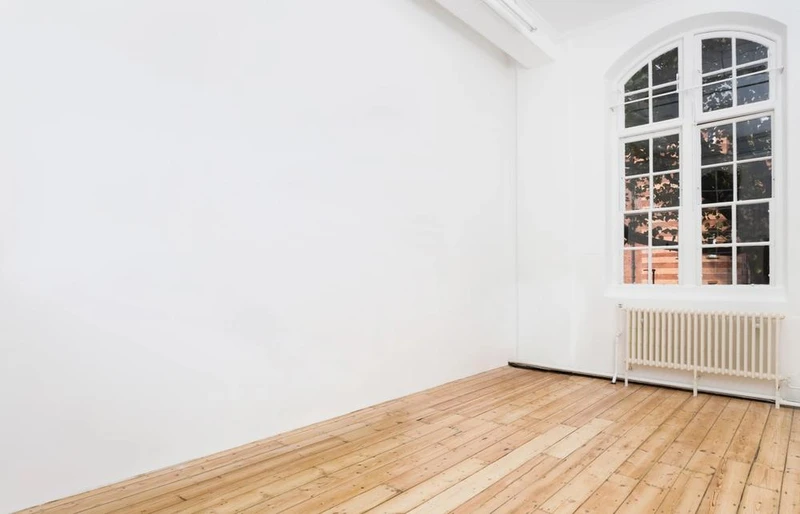 A room featuring a white wall and wood flooring, with a door and window visible. The interior has a clean design with elements of hardwood and plywood. | 2025 Maureen Paley: Studio M