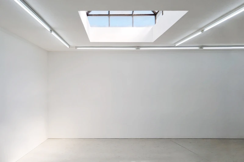 A bright room featuring a skylight, with white walls and a whiteboard. Fluorescent lamps illuminate the space, highlighting text and artistic designs on the surfaces. The ceiling is visible, enhancing the open feeling of the indoor environment. | 2025 Maureen Paley