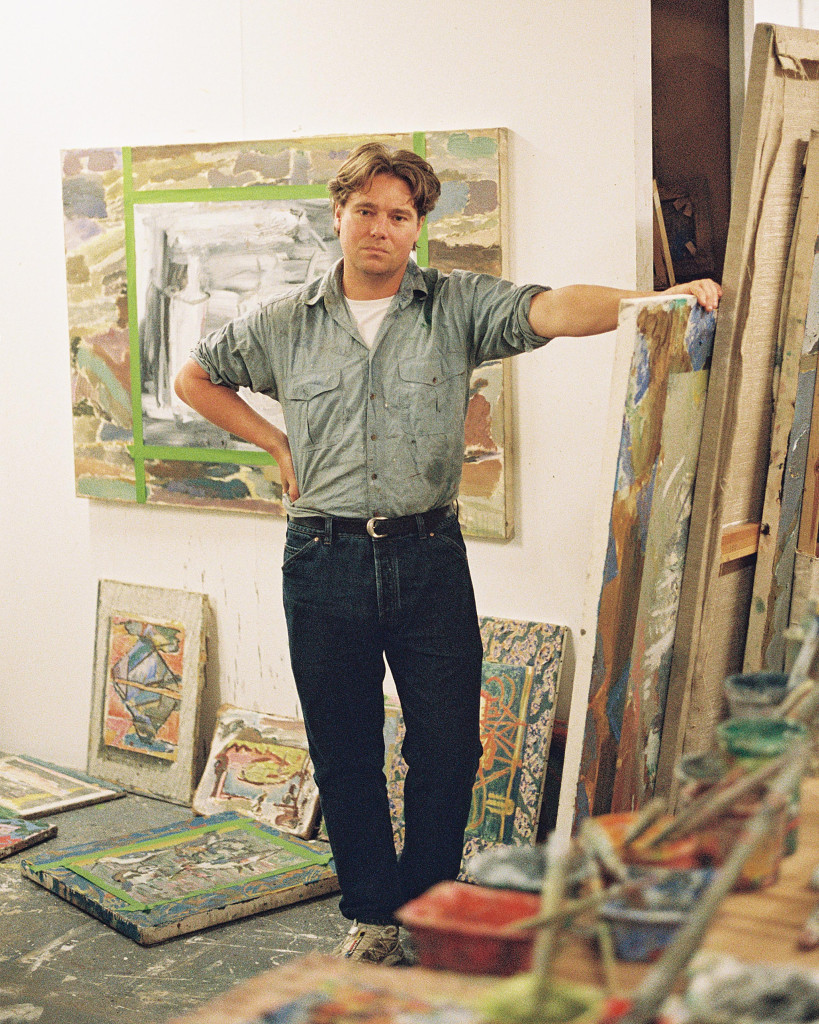 A man wearing jeans and a casual top stands in front of a painting framed on the wall indoors.