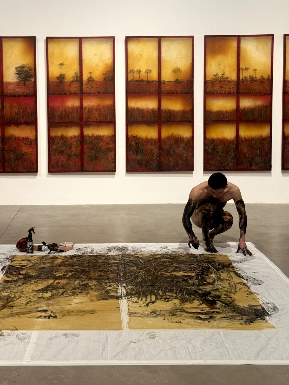 A man is standing in an art gallery, focused on painting a large canvas hung on the wall. The indoor space is filled with various artworks and has a creative ambiance.