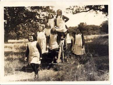 A group of girls standing together on a wooden swing, surrounded by green grass and trees in an outdoor setting.