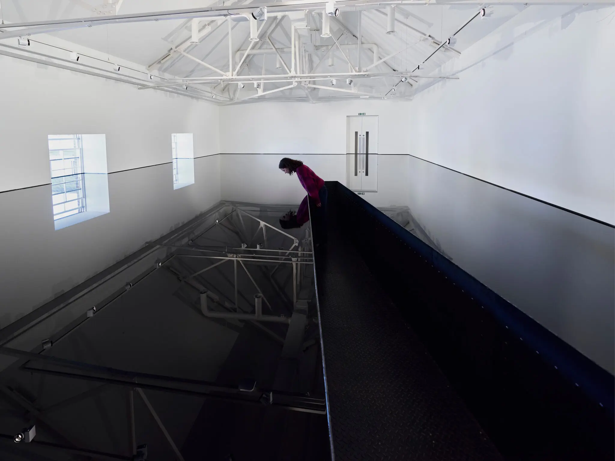 A person standing on a black railing indoors, surrounded by a white wall and art displayed on a whiteboard, with visible ceiling and floor details.