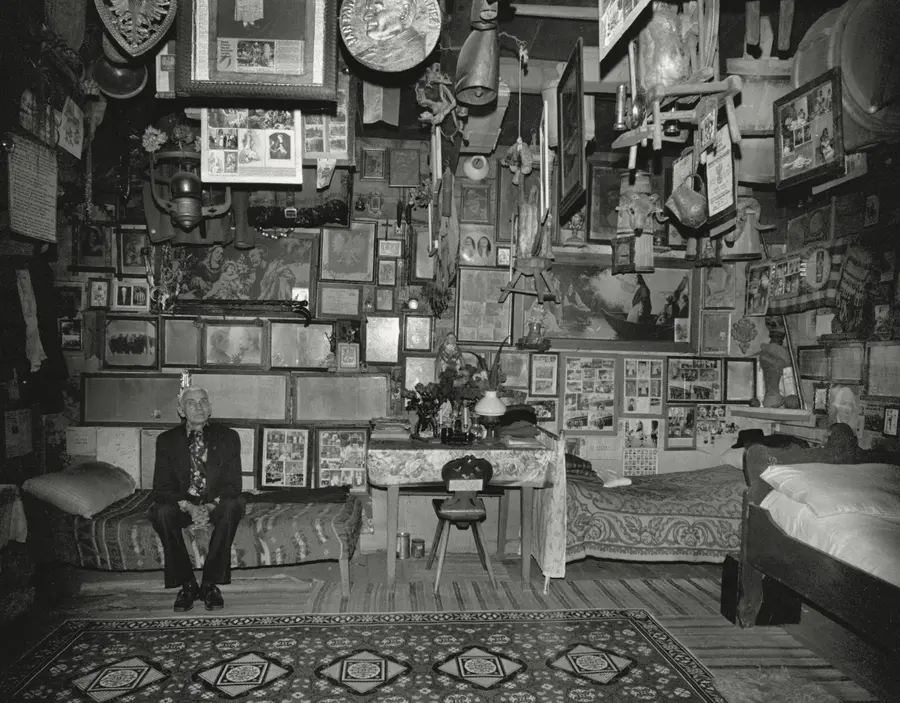 A man is sitting on a couch in a room filled with pictures on the walls and a table nearby. The image is in black and white, featuring elements of furniture, a clock, and an indoor setting.