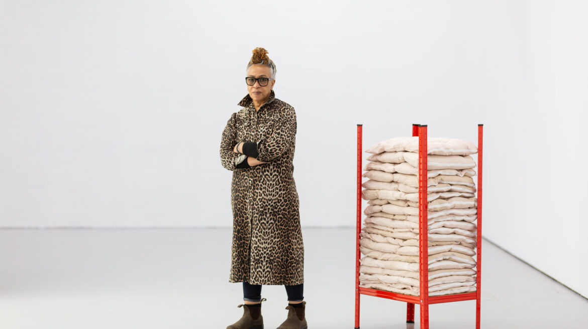 A woman wearing a stylish coat stands next to a tall stack of sacks, showcasing a blend of fashion and casual elements in her attire.