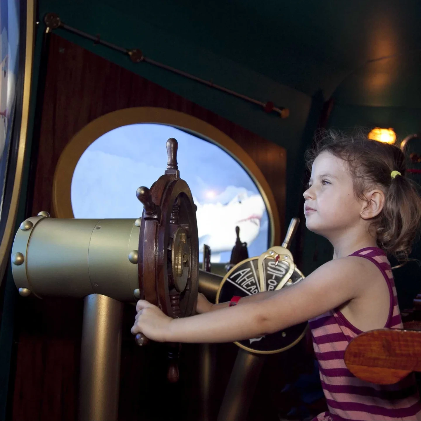 A young girl outdoors, holding a steering wheel, wearing casual clothing.