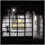 A window featuring a painting, surrounded by the interior of a building. Outside, a tree is visible under the night sky with soft light illuminating the scene.
