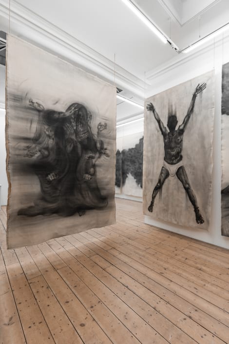 A collection of paintings displayed on a wall in an indoor art gallery, featuring various artworks with a decorative ceiling and room floor visible.