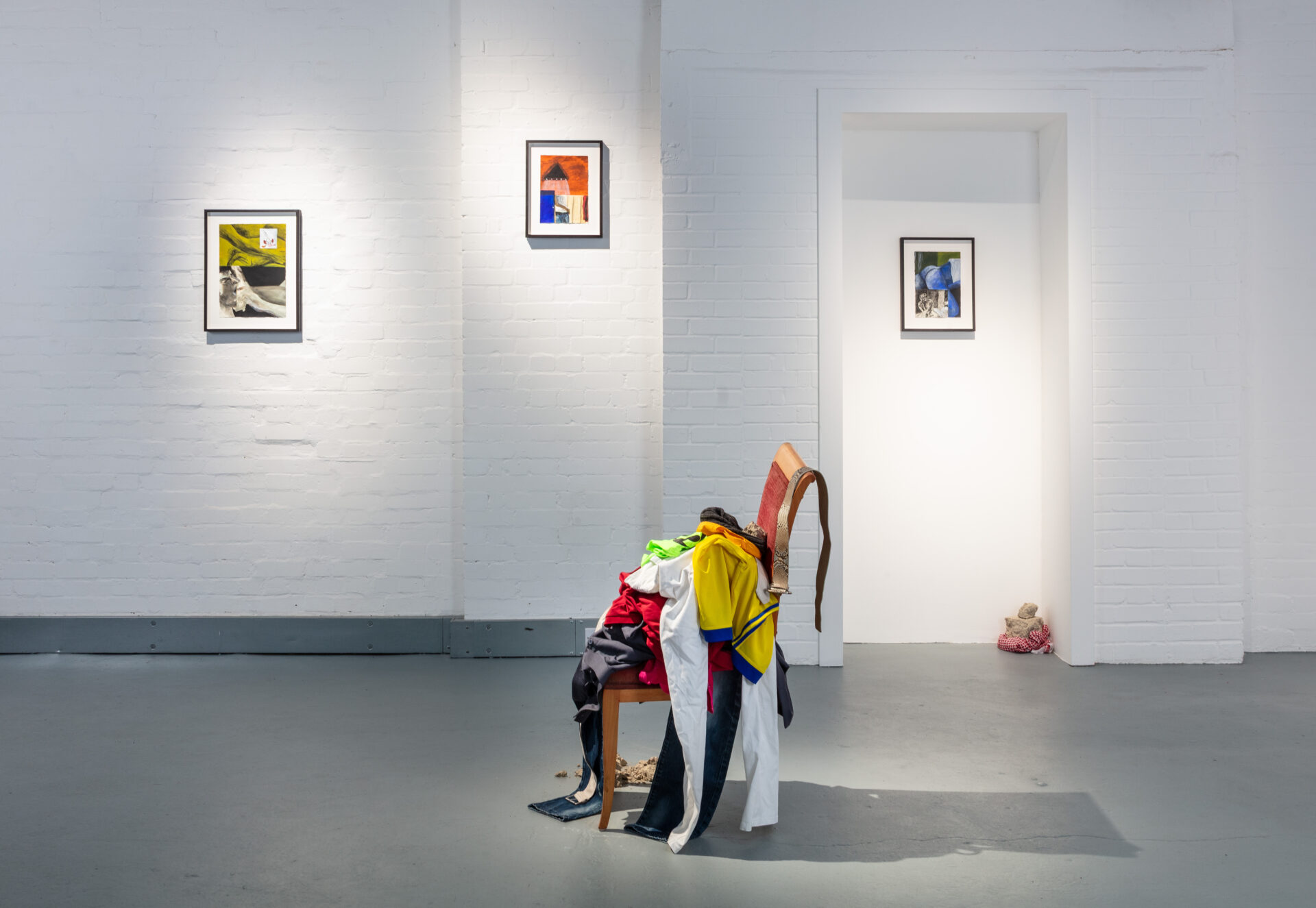 An art gallery featuring two framed artworks on the walls, with a chair overloaded with colorful clothes in the foreground.