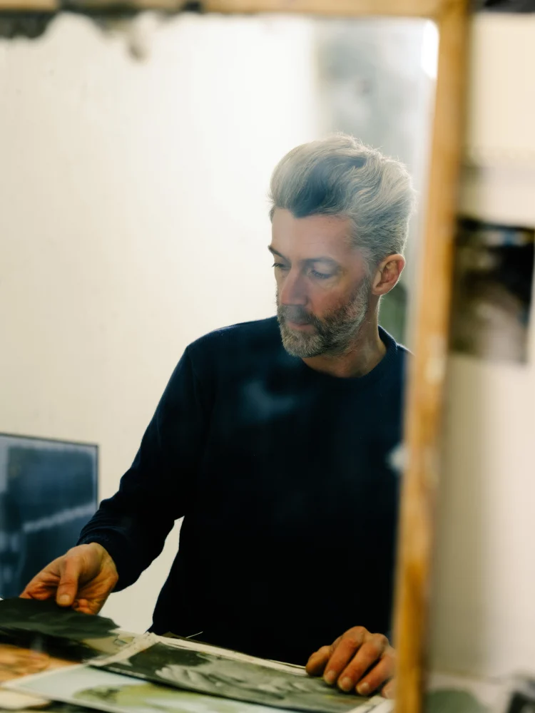 A man standing indoors, looking intently at a record while surrounded by a wall and a computer with a keyboard.