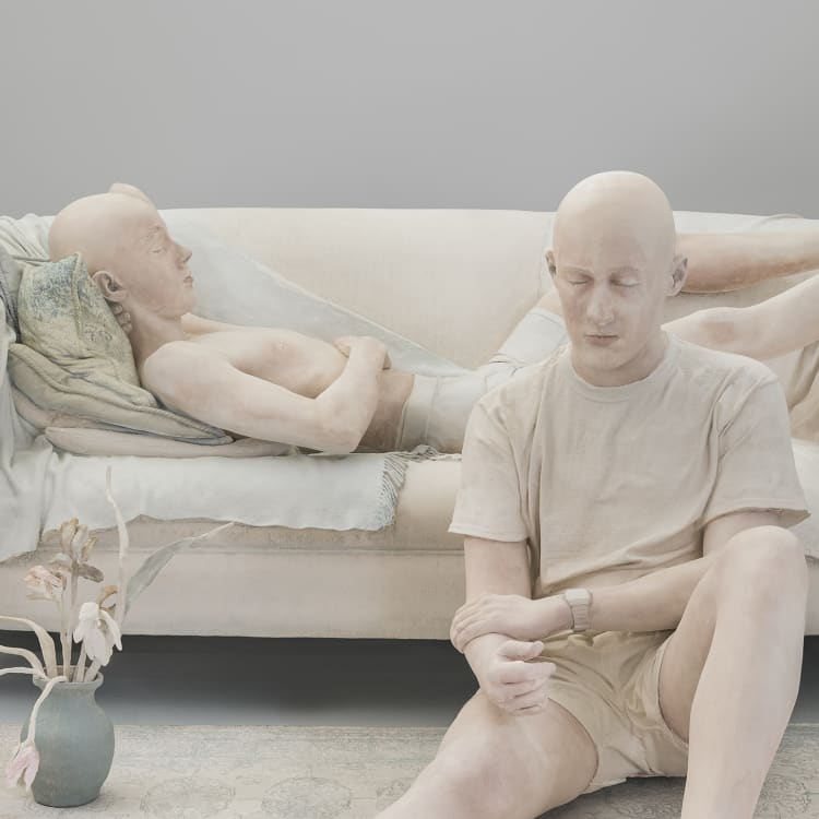 A minimalist living room scene with two individuals in neutral-colored clothing on a light sofa, surrounded by soft decor and a flower vase.