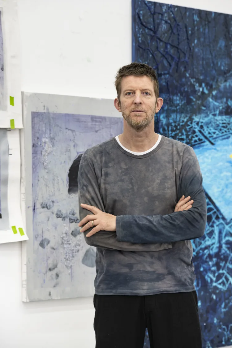 A man is standing indoors with his arms crossed, wearing trousers and a stylish shirt. He has a focused expression on his face, and there is a wall in the background.